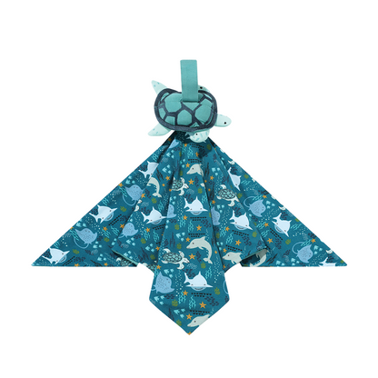 the "ocean friends lovey" is a blanket with the "ocean friends" print and a turtle perched on the top of it. the "ocean friends" print is a combination of dolphins, stingrays, fish, starfish, coral, bubbles, and sharks, all spread out on a deep sea blue background. 