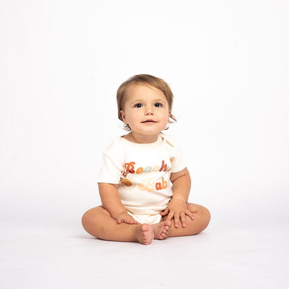 a baby sitting on the floor in the "beach babe" cotton onesie. the word 'beach babe' is spelt out in a groovy font with a sun, starfish, and shell surrounding it. 
