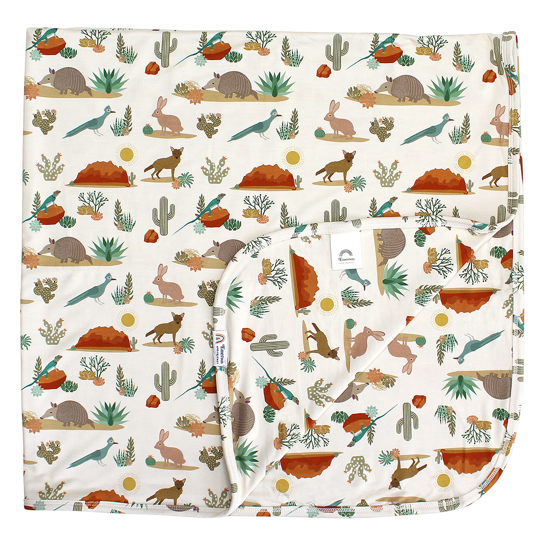 the "desert friends" bamboo blanket. the "desert friends" print is a scattered pattern of armadillo, cacti, other desert plants, rocks, desert foxes, lizards, and desert birds. this is all on a beige background. 