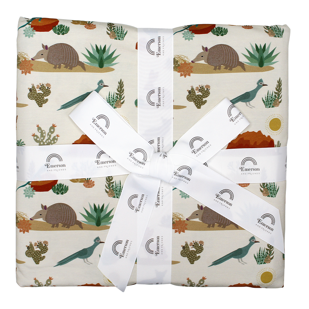 the "desert friends" twin sized bamboo blanket. the "desert friends" print is a scattered pattern of armadillo, cacti, other desert plants, rocks, desert foxes, lizards, and desert birds. this is all on a beige background. 