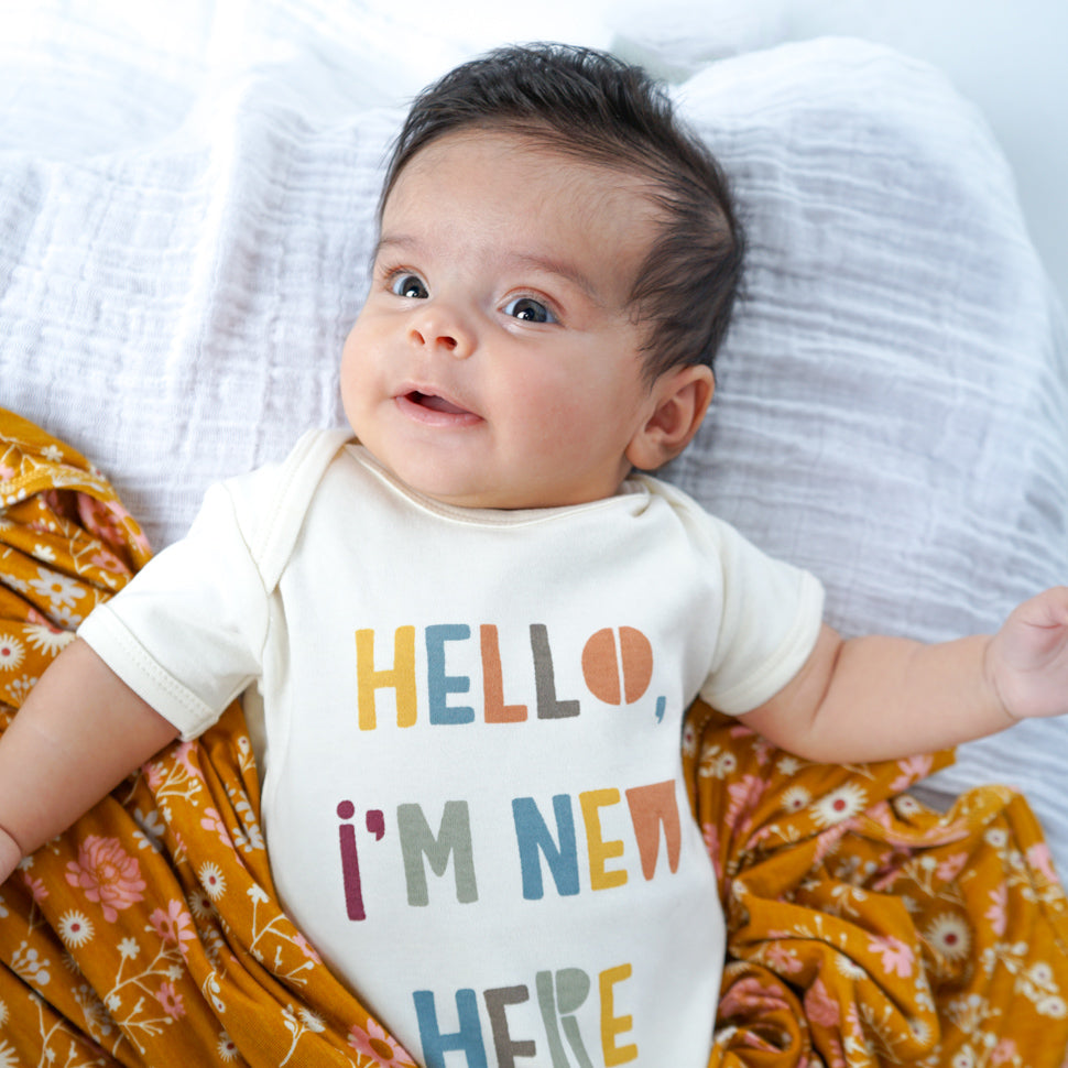 baby lays on a white pillow. the baby is half covered in the "mustard floral" blanket. the baby wears the "hello, im new here" onesie. the "mustard floral" print are a bunch of white and pink floral flowers on a mustard yellow background. 