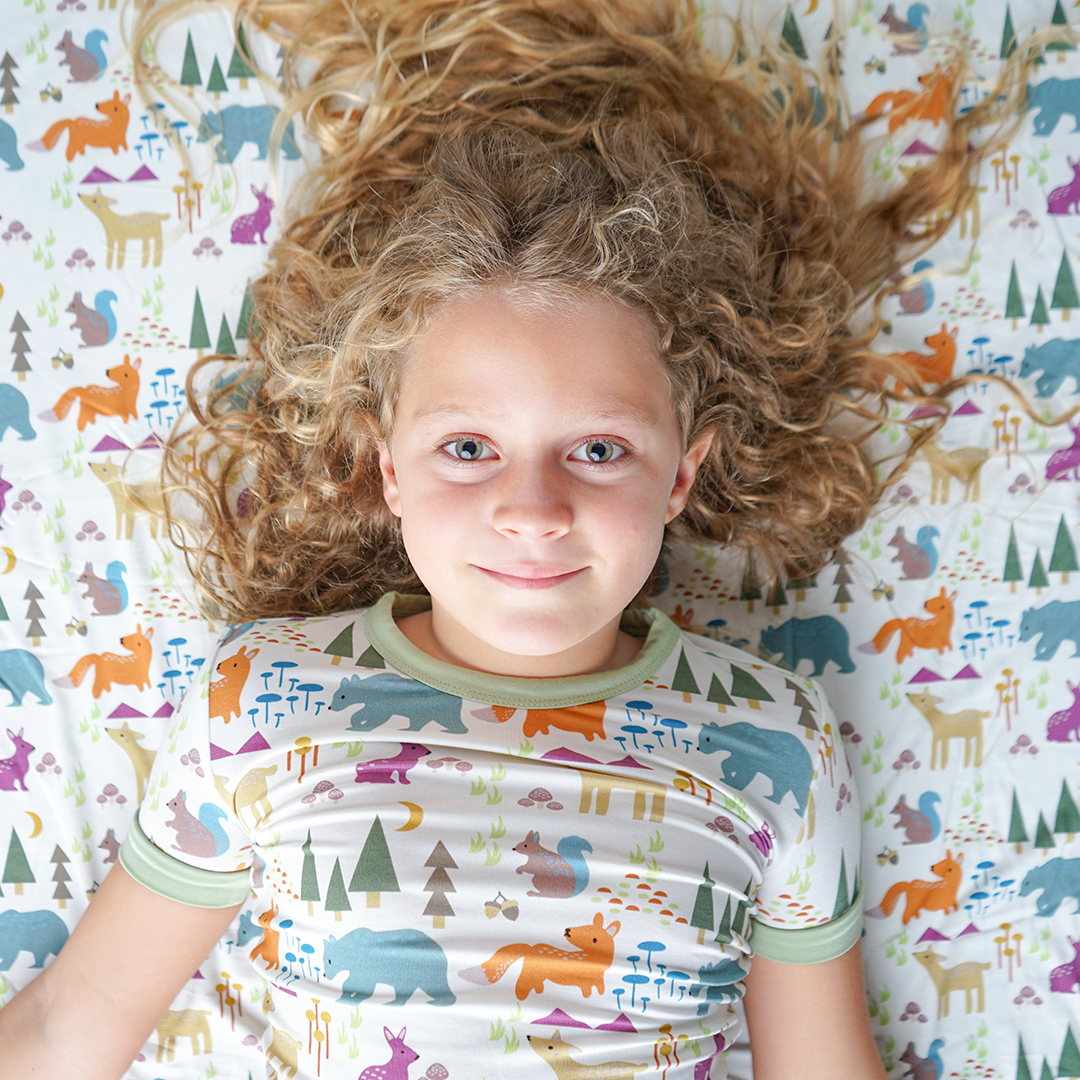 young girl lays on the "forest friends" bamboo blanket. she is wearing the "forest friends" matching pajama set. the "forest friends" print has a mix of red foxes, brown deer, blue bears, purple bunnies, trees, grass, mushrooms, acorns, moons, and other colored dots scattered over a white background. 