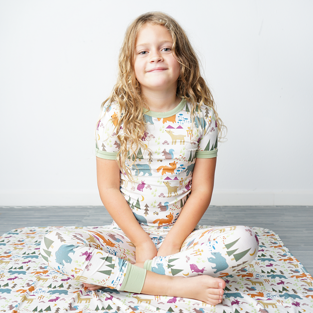 young girl sits criss-cross with her hands in her lap. she sits on the "forest friends" bamboo blanket. she wears the "forest friends" matching pajama set. the "forest friends" print has a mix of red foxes, brown deer, blue bears, purple bunnies, trees, grass, mushrooms, acorns, moons, and other colored dots scattered over a white background. 