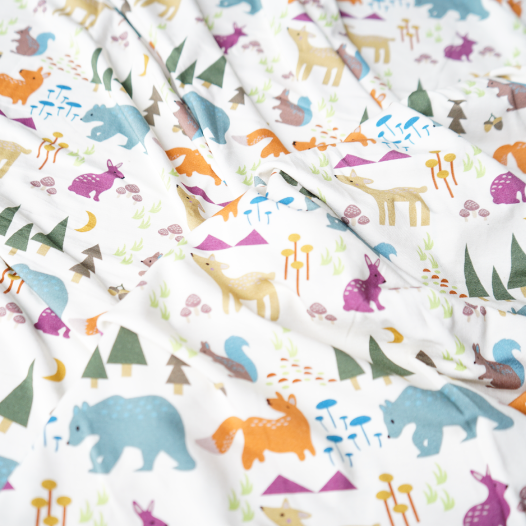 a zoomed in photo of the print of the"forest friends" blanket. the "forest friends" print has a mix of red foxes, brown deer, blue bears, purple bunnies, trees, grass, mushrooms, acorns, moons, and other colored dots scattered over a white background. 