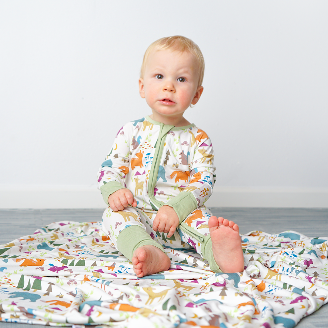 a young baby sits on the "forest friends" bamboo blanket. the baby wears the "forest friends" convertible. the "forest friends" print has a mix of red foxes, brown deer, blue bears, purple bunnies, trees, grass, mushrooms, acorns, moons, and other colored dots scattered over a white background. 