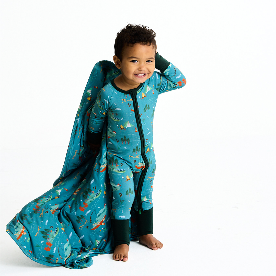 young boy smiles and holds the "jump in the lake" blanket. he wears the "jump in the lake" convertible. the "jump in the lake" pattern is a print to capture the day in the life on a lake. you can fine fishing poles, tents, camp fires, adirondack chairs, marsh mellows, ores, canoes, forest trees, and different types of fish and fishing gear.