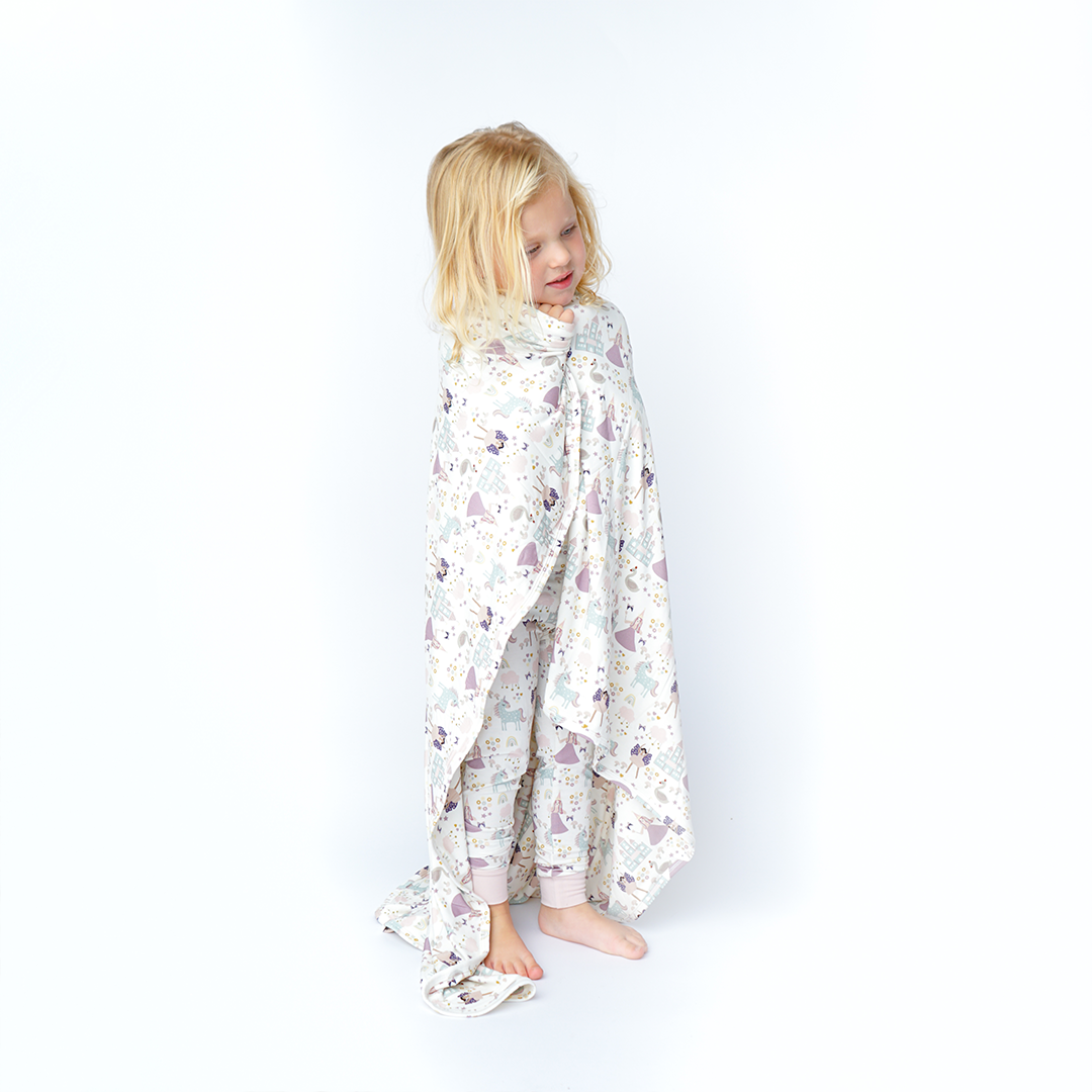 a young girl wraps herself in the "once upon a time" bamboo blanket. the "once upon a time" print is a mix of pinks, purples, whites, blues, and yellows. you can see fairy princess and regular princess, unicorns, swans, and castles. theres also hearts, stars and sparkles scattered around the print. 