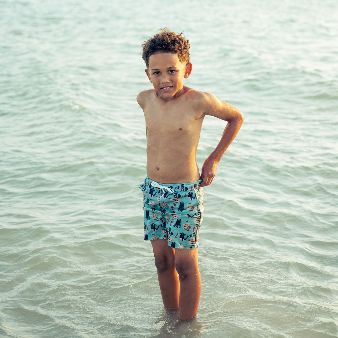 a boy poses in the ocean. he is wearing the "pirate's life" swim trunks. the "pirate's life" print is a mix of octopus, whales, and seagulls all wearing pirate hats. there are also anchors, pirate ships, treasure maps, and starfish mixed into the pattern. this is all displayed on a teal background. 