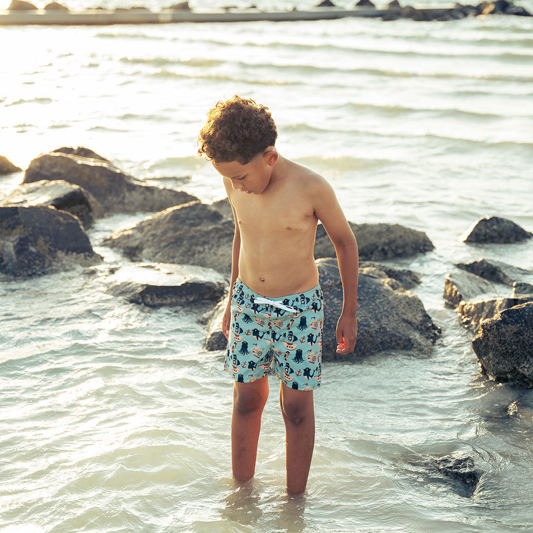 a boy stands in the water with a dozen rocks behind him. he is wearing the "pirate's life" swim trunks. the "pirate's life" print is a mix of octopus, whales, and seagulls all wearing pirate hats. there are also anchors, pirate ships, treasure maps, and starfish mixed into the pattern. this is all displayed on a teal background. 