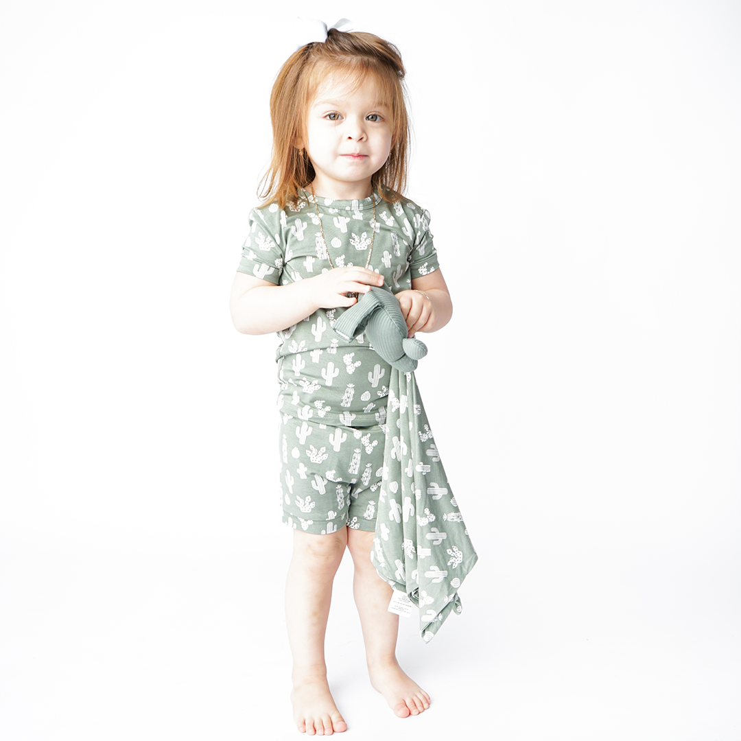 girl stands and holds the "stay sharp" lovey. she is wearing the "stay sharp" matching pajama set. the "stay sharp" print is a variety of different white cacti on a greyish/green background. 