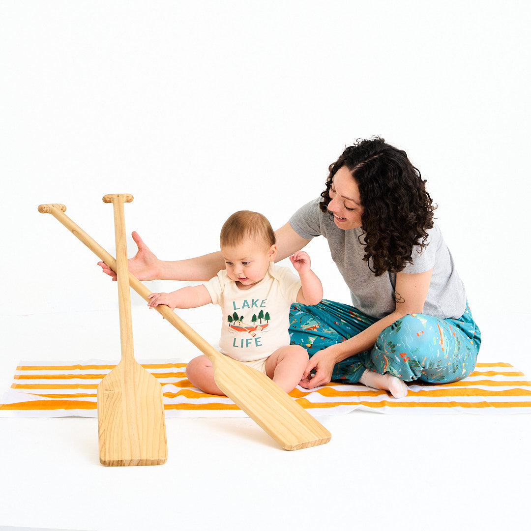 mom and baby sit on a orange and white striped towel. they play with canoe ores. the baby is in the "lake life" cotton onesie. this design says the phrase "lake life" and has a canoe, ores, oak trees, and some lake waves. 