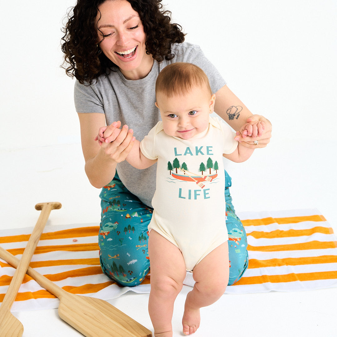 mom sits on the floor and hold her baby's hands up. the baby stands and wears the "lake life" cotton onesie. the mom is in the "jump in the lake" relaxed pants. they sit on a orange and white striped towel. this design says the phrase "lake life" and has a canoe, ores, oak trees, and some lake waves. 