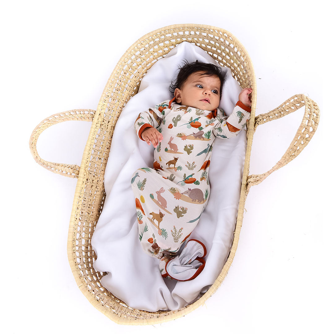  a small baby lays in a basket with a white, fluffy blanket. the baby is in the "desert friends" gown. the gown is tied in a knot at the bottom. the "desert friends" print is a scattered pattern of armadillo, cacti, other desert plants, rocks, desert foxes, lizards, and desert birds. this is all on a beige background. 