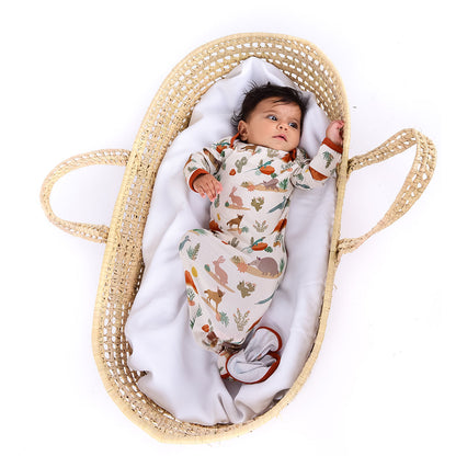  a small baby lays in a basket with a white, fluffy blanket. the baby is in the "desert friends" gown. the gown is tied in a knot at the bottom. the "desert friends" print is a scattered pattern of armadillo, cacti, other desert plants, rocks, desert foxes, lizards, and desert birds. this is all on a beige background. 