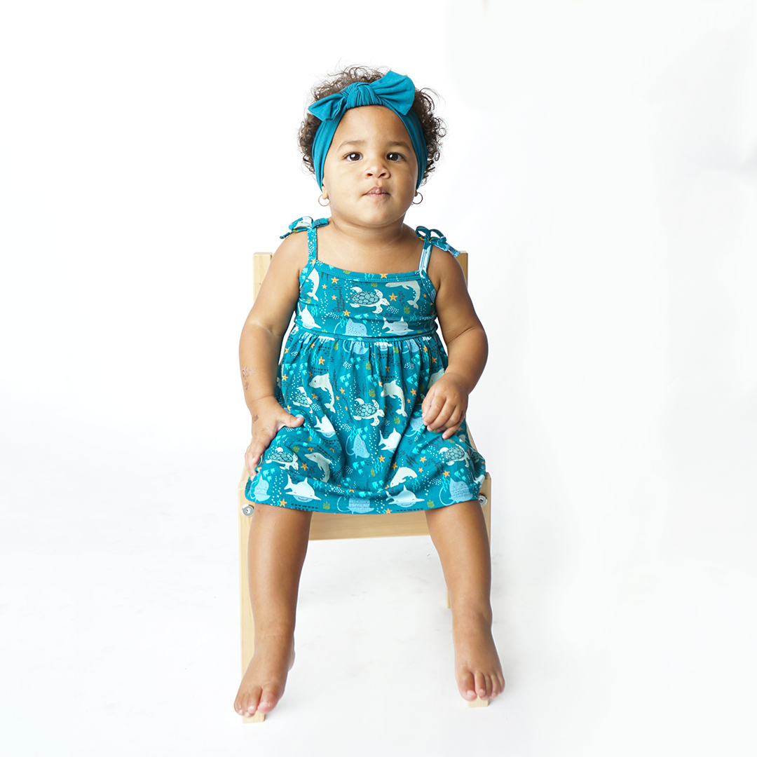 baby sits in a wooden chair in her "ocean friends" sundress and the "ocean blue" headband. the "ocean friends" print is a combination of dolphins, stingrays, fish, starfish, coral, bubbles, and sharks, all spread out on a deep sea blue background. 