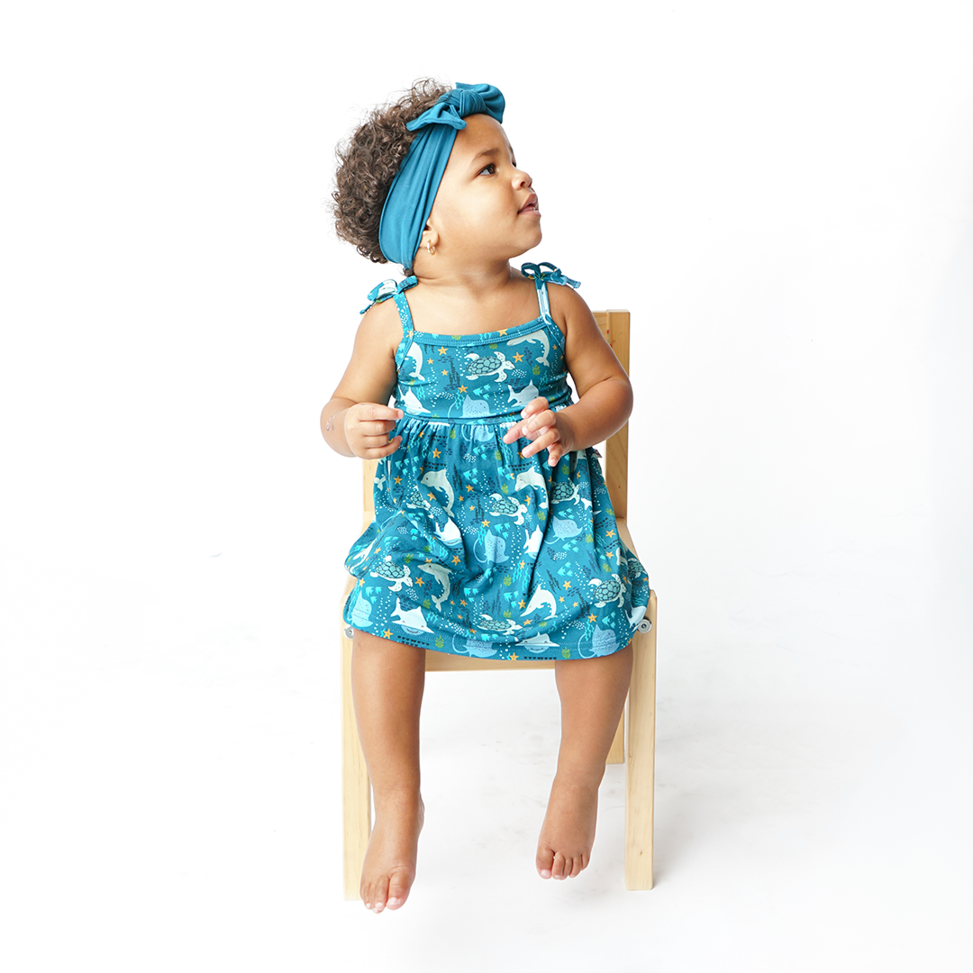 a baby girl sits in a wooden chair, looking away from the camera. she is wearing the "ocean friends" sundress and the "ocean blue" headband. the "ocean friends" print is a combination of dolphins, stingrays, fish, starfish, coral, bubbles, and sharks, all spread out on a deep sea blue background. 