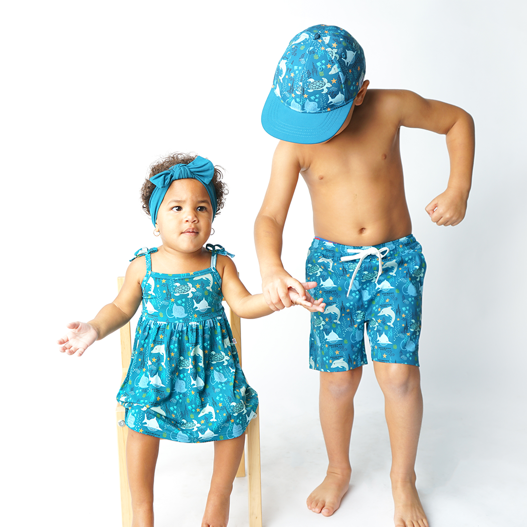 baby girl sits on a wooden chair wearing the "ocean friends" sundress and the "ocean blue" headbands. she holds her brothers hand. the brother stands facing the floor in his "ocean friends" swim trunks and snapback hat. v