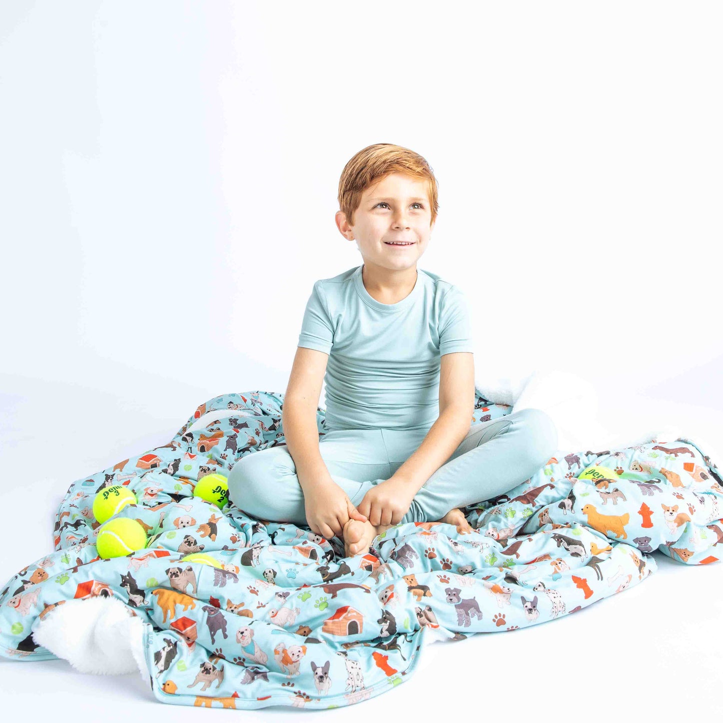 young boy sits on the "dogs bark" fleece blanket. there are tennis balls scattered around him. the "dogs bark" print is a mix of various types of dogs, dog houses, dog bones, paw prints, dog food, and tennis balls scattered around a light blue background.