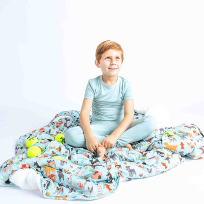 young boy sits on the "dogs bark" fleece blanket. there are tennis balls scattered around him. the "dogs bark" print is a mix of various types of dogs, dog houses, dog bones, paw prints, dog food, and tennis balls scattered around a light blue background.