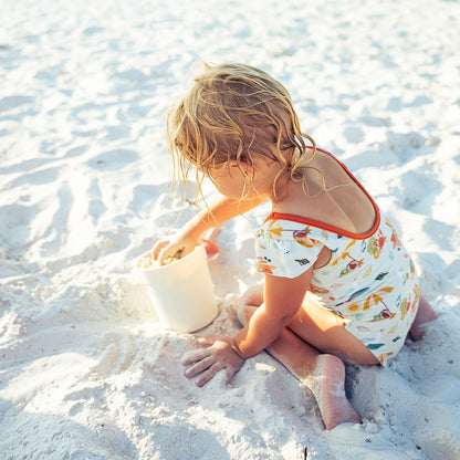 young girl plays in the sand with a toy bucket. she is wearing the "beach days" one-piece swim suit. the "beach days" print is a scattered print of beach umbrellas, beach balls, sand buckets and shovels, coral, shells, beach chairs, seagulls, flipflops, and sunglass mixed in a orderly and creative way. 