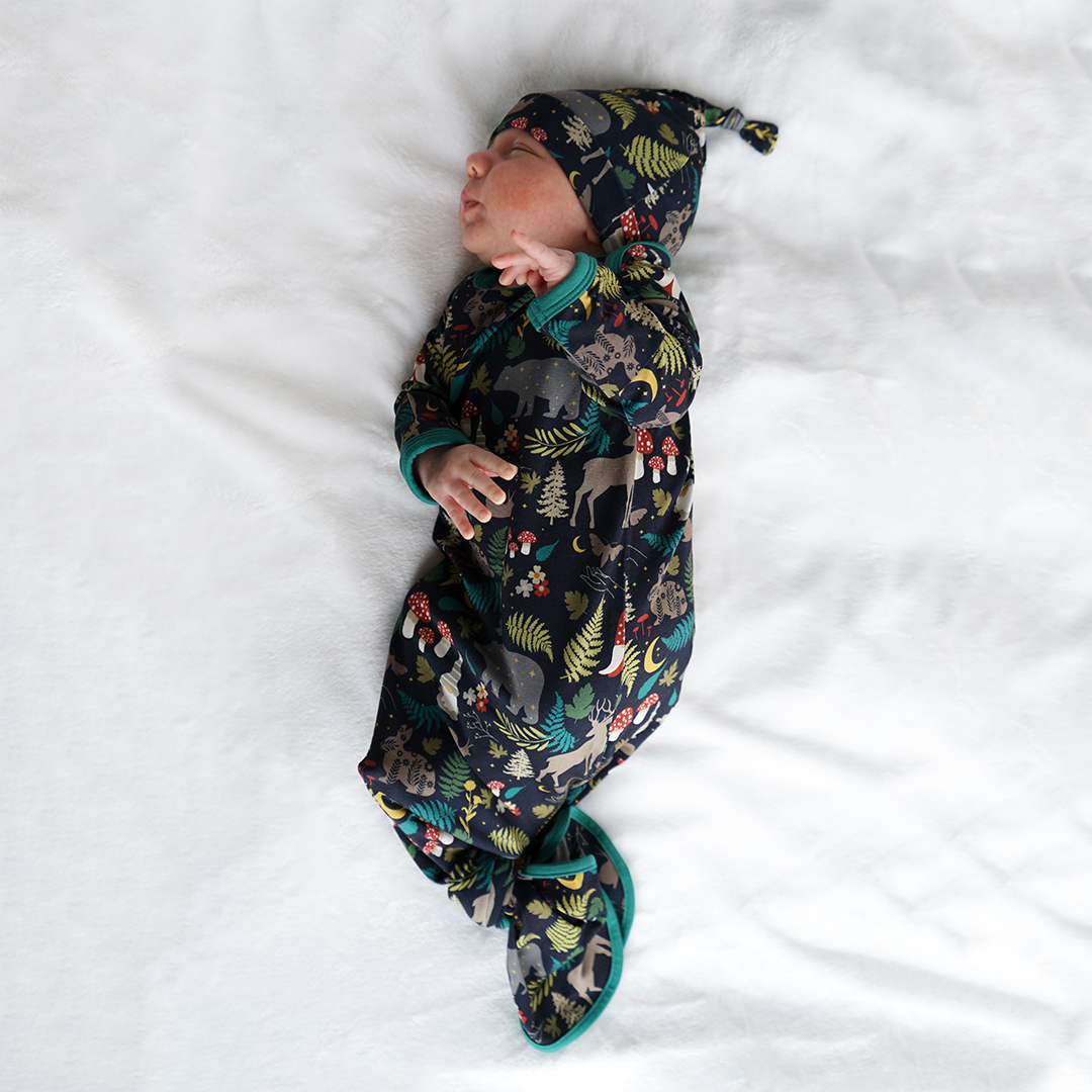 a infant baby is seen laying on his side on a white blanket. the baby is in the "night forest" gown and hat set. the "night forest" print is a night time themed design. you can see an array of forest animals ranging from, deer, bears, bunnies, birds, forest trees and leaves, flowers, and mushrooms. there are also starts and moons scattered around to enhance the night time atmosphere. 