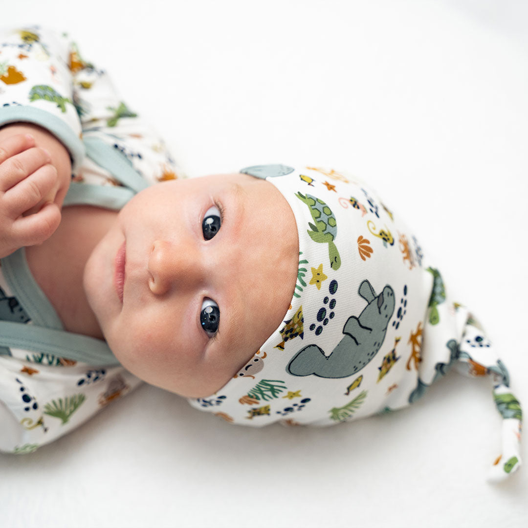 a baby stares into the camera in their "manatee" gown and hat set. the print "manatee" unites the florida sea life in a majestic print. there is a variation of florida manatees, yellow and pink sea horses, yellow and pink starfish, bubbles, turtles, and coral patterned across a white background