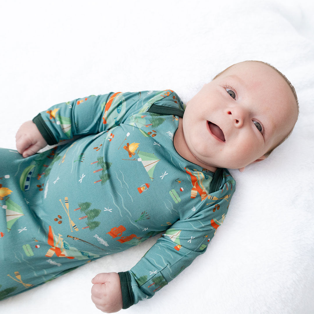 infant baby smiles at the camera. the baby wears the "jump in the lake" gown and hat set. the "jump in the lake" pattern is a print to capture the day in the life on a lake. you can fine fishing poles, tents, camp fires, adirondack chairs, marsh mellows, ores, canoes, forest trees, and different types of fish and fishing gear.