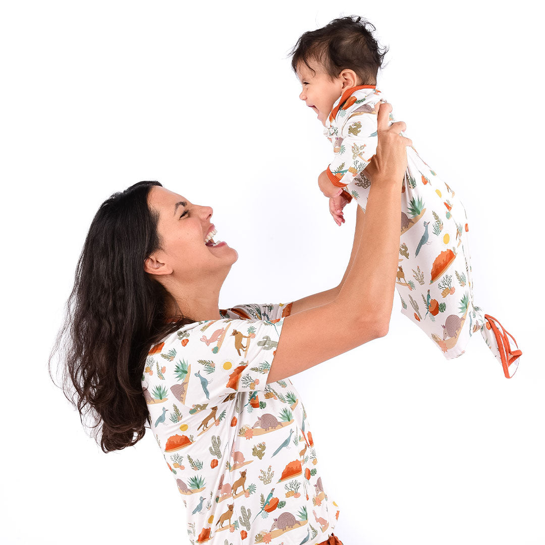 mom smiles and laughs while she raises her baby in the air. the mom and baby are both wearing matching "desert friends" prints. the mom is in the women's top and the babyis in the gown. the "desert friends" print is a scattered pattern of armadillo, cacti, other desert plants, rocks, desert foxes, lizards, and desert birds. this is all on a beige background. 