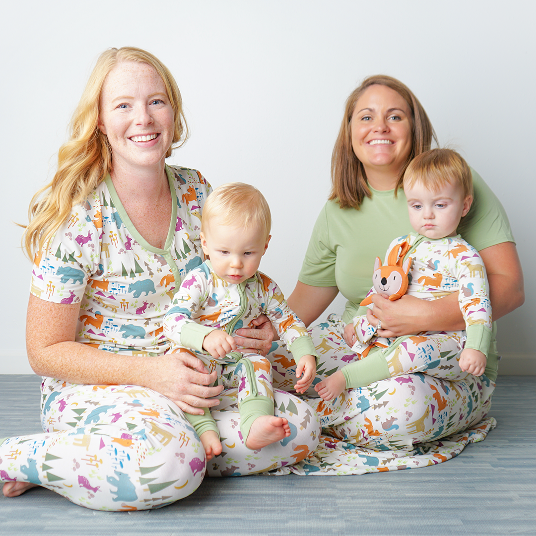 2 moms sit on the floor with their babies in their lap, all in "forest friends" apparel. the moms are wearing the women's joggers and women's top. the babies are in the convertibles. one of the babies holds a lovey. the "forest friends" print has a mix of red foxes, brown deer, blue bears, purple bunnies, trees, grass, mushrooms, acorns, moons, and other colored dots scattered over a white background. 