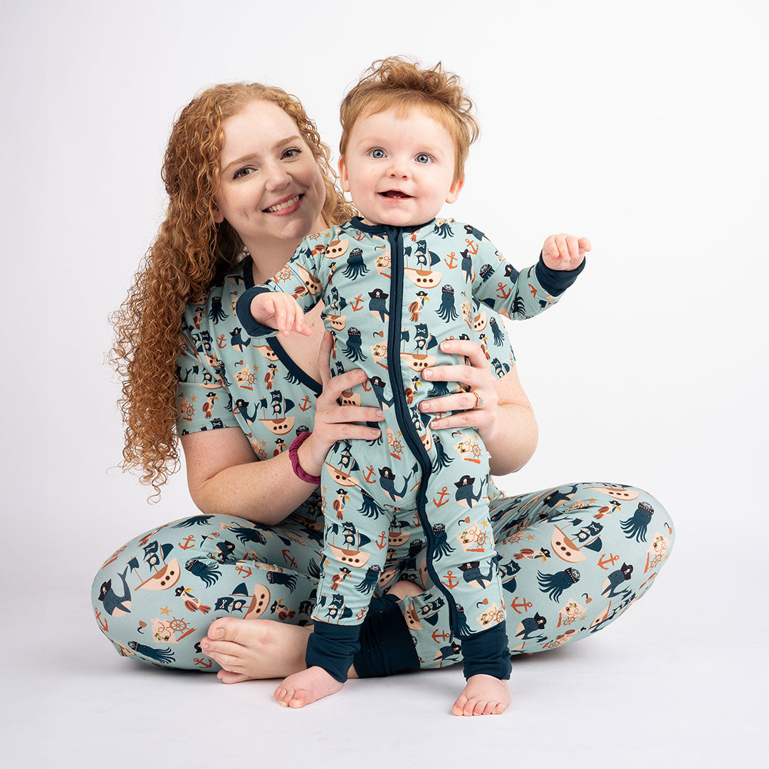 a mom and baby smile at the camera together. the mom sits on the floor, she is wearing the "pirate's life" women's joggers and the women's top. the baby wears the "pirate's life" convertible. the "pirate's life" print is a mix of octopus, whales, and seagulls all wearing pirate hats. there are also anchors, pirate ships, treasure maps, and starfish mixed into the pattern. this is all displayed on a teal background. 