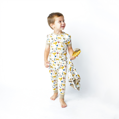 a boy walks and looks at something off camera. he is wearing the "jungle friends" matching pajama set and holds the lion lovey. the "jungle friends" print is a mix of jungle animals that include giraffes, lions, cheetahs, sloths, snakes, and forest plants. 