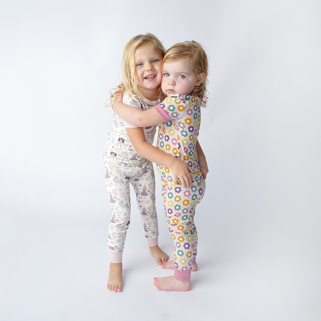 2 sisters hug. they are both wearing 2-piece matching pajama sets. the oldest wears the "once upon a time" set. the "once upon a time" print is a mix of pinks, purples, whites, blues, and yellows. you can see fairy princess and regular princess, unicorns, swans, and castles. theres also hearts, stars and sparkles scattered around the print. 