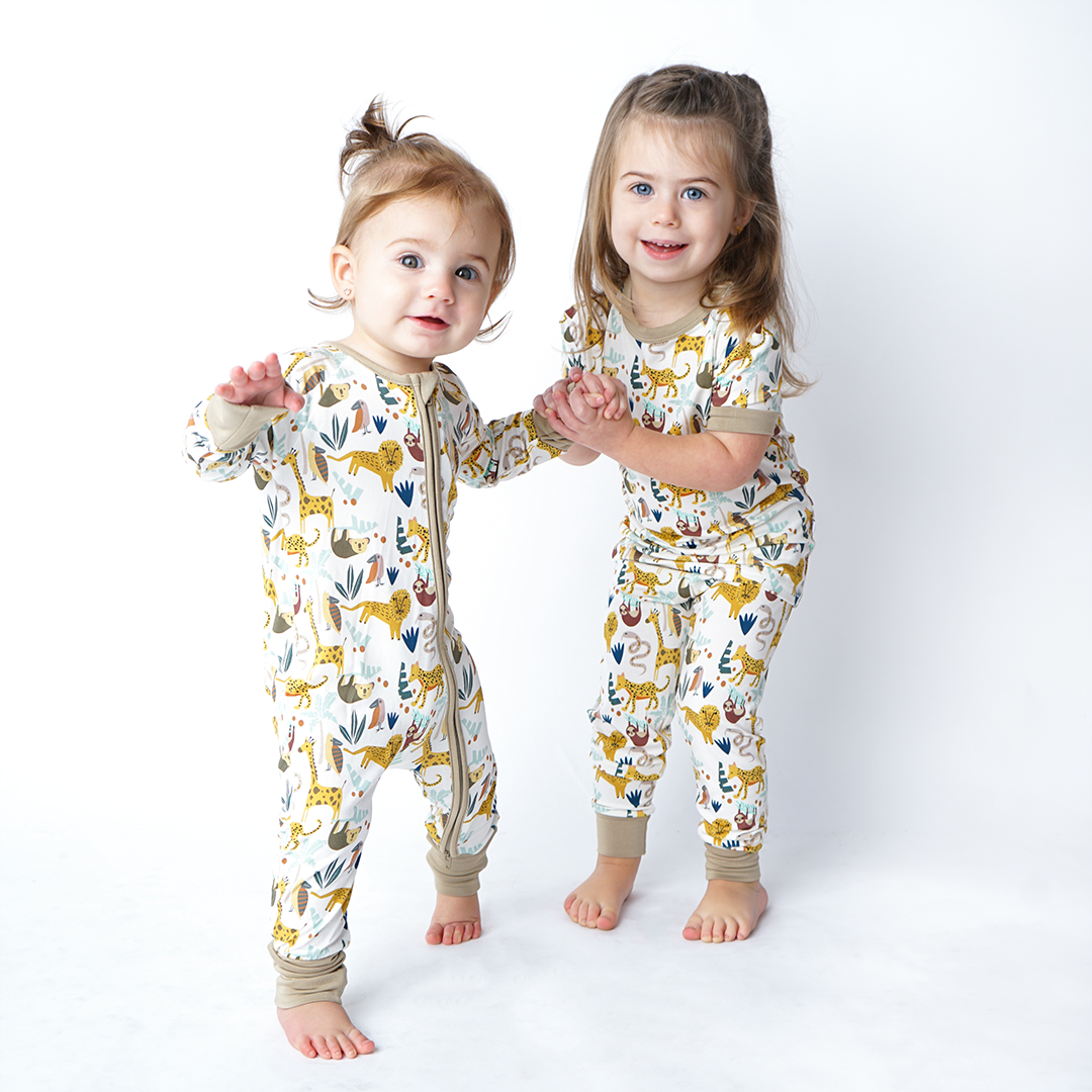 2 girls hold hands and wear the "jungle friends" pajamas. the youngest is in the convertible. the older one is in the matching pajama ser. the "jungle friends" print is a mix of jungle animals that include giraffes, lions, cheetahs, sloths, snakes, and forest plants. 