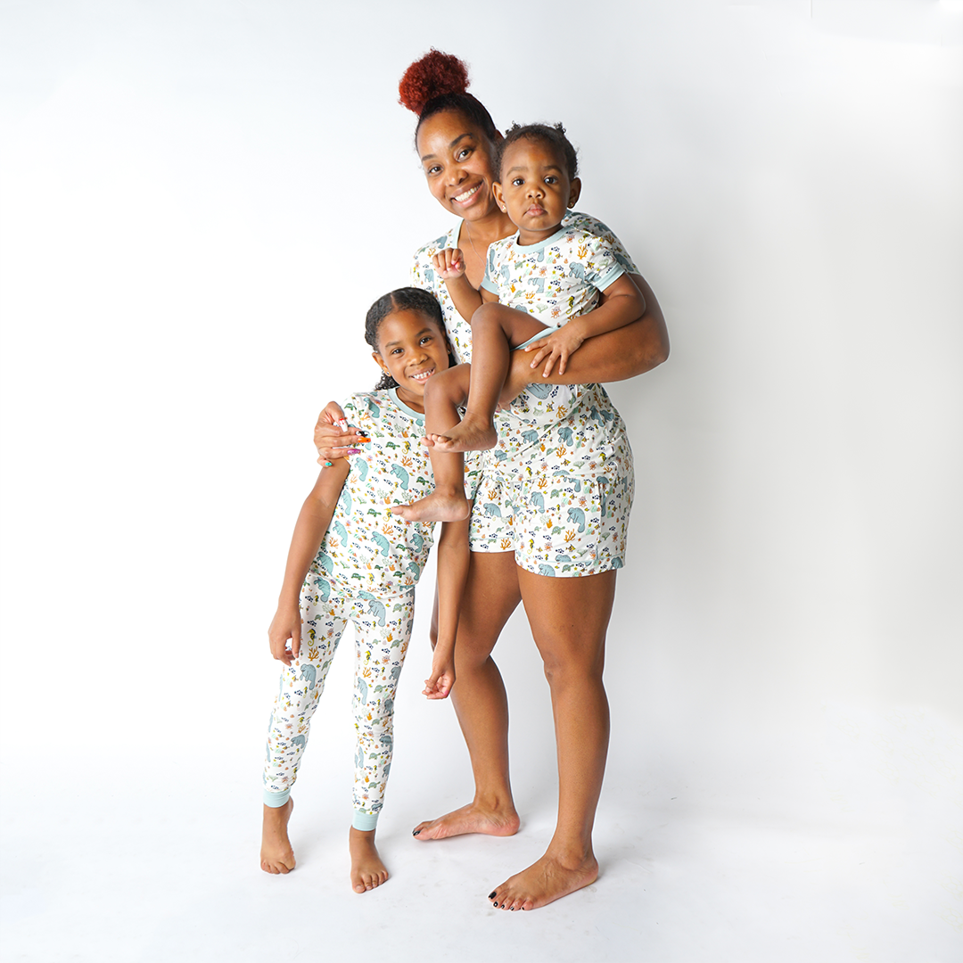 mom of two stands with her kids. she holds one of them in her arms who is wearing the "manatee" shortie. the other child stands next to to the mom in her "manatee" 2-piece matching set. the mom embraces both kids in her "manatee" women's top and women's shorts. the print "manatee" unites the florida sea life in a majestic print. there is a variation of florida manatees, yellow and pink sea horses, yellow and pink starfish, bubbles, turtles, and coral patterned across a white background 