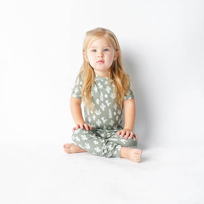 a little girl sits criss-cross on the floor and stares at the camera. she is wearing the 2-piece "stay sharp" matching pajama set. the "stay sharp" print is a variety of different white cacti on a greyish/green background. 