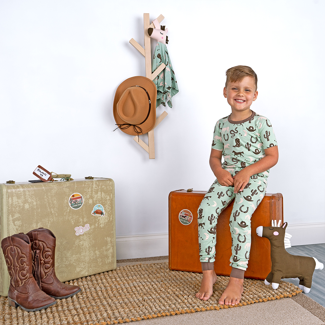 a young boy sits on top of a western looking suit case. he wears the "giddy up" matching pajama set. there is a western set up centered around him that includes another suit case, cowboy boots, a cowboy hat, and the "giddy up" lovey. the "giddy up" print is a western look at horses, cowboy hats, boots, horse rings, cacti, moons and stars. colors on this print include green, brown, and white. 