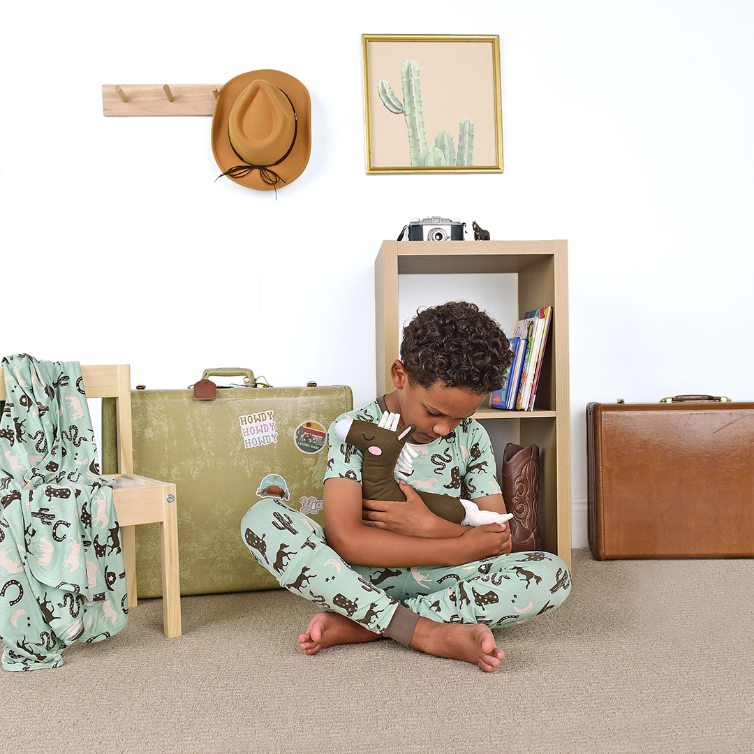 young boy sits in a western looking room. he wears the "giddy up" matching pajama set. he is holding the lovey in his arms. the "giddy up" print is a western look at horses, cowboy hats, boots, horse rings, cacti, moons and stars. colors on this print include green, brown, and white. 