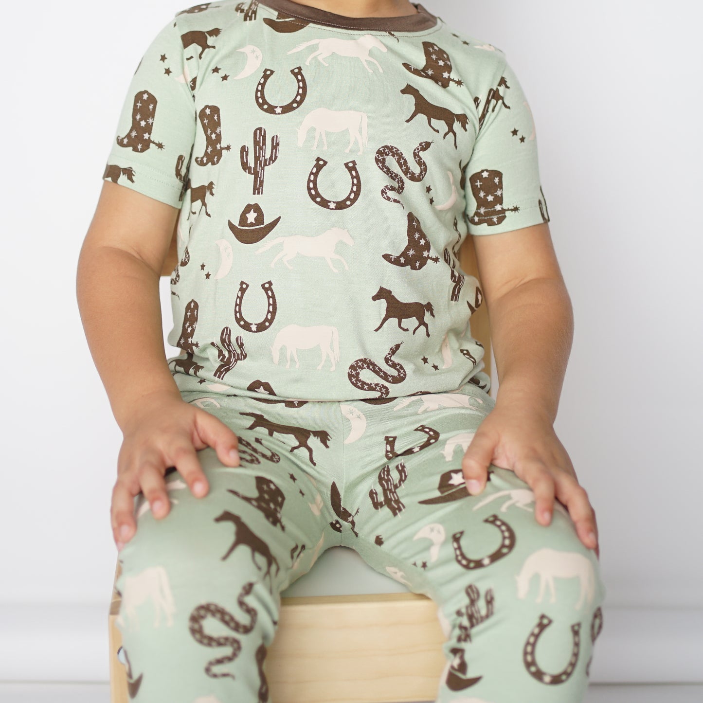 close up of a child sitting on a wooden chair. the child wears the "giddy up" pajama set. the "giddy up" print is a western look at horses, cowboy hats, boots, horse rings, cacti, moons and stars. colors on this print include green, brown, and white. 