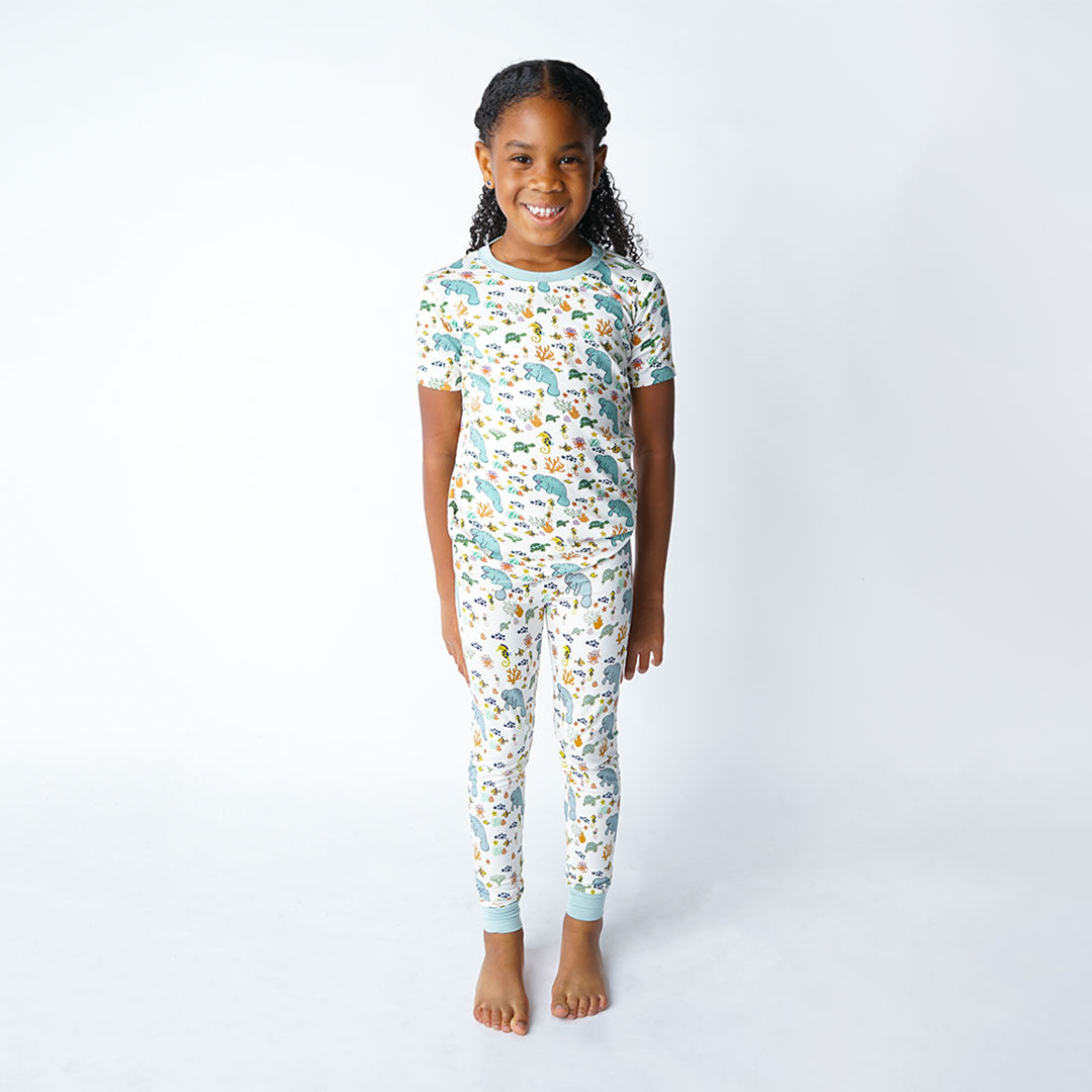 girl poses with her arms at her side wearing the 2-piece matching pajama set. the print "manatee" unites the florida sea life in a majestic print. there is a variation of florida manatees, yellow and pink sea horses, yellow and pink starfish, bubbles, turtles, and coral patterned across a white background
