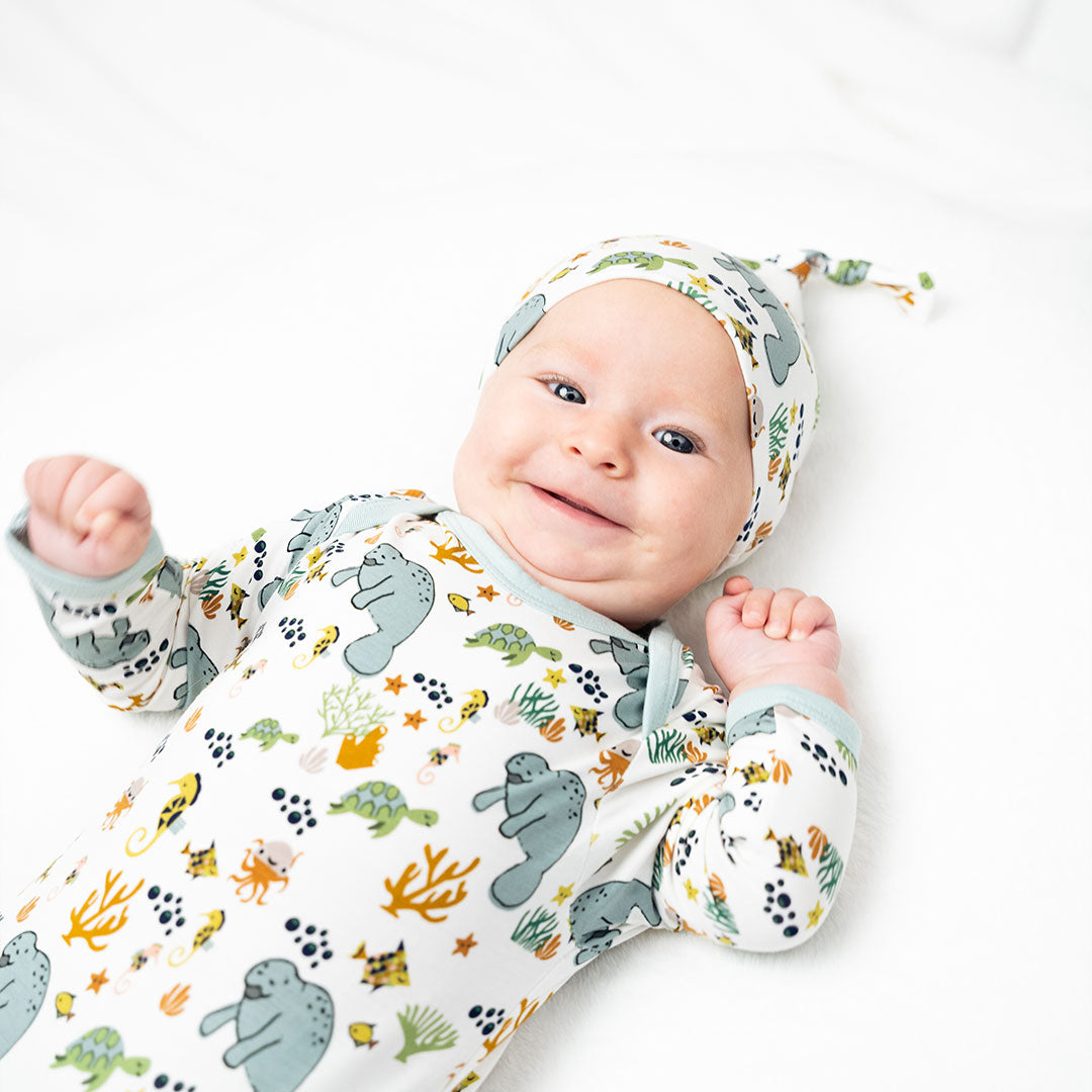 a happy, smiling baby lays on their back with the "manatee" gown and hat set. the print "manatee" unites the florida sea life in a majestic print. there is a variation of florida manatees, yellow and pink sea horses, yellow and pink starfish, bubbles, turtles, and coral patterned across a white background