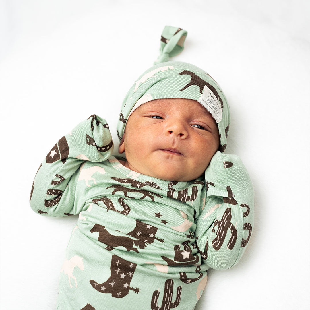 small infant in the "giddy up" gown and hat. the "giddy up" print is a western look at horses, cowboy hats, boots, horse rings, cacti, moons and stars. colors on this print include green, brown, and white. 