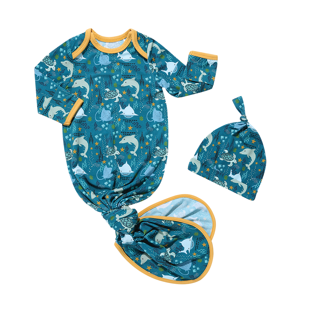 the "ocean friends" bamboo newborn gown and hat baby set. the gown ties at the bottom. the "ocean friends" print is a combination of dolphins, stingrays, fish, starfish, coral, bubbles, and sharks, all spread out on a deep sea blue background. 