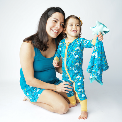 mom and child are on the floor in their "ocean friends" apparel. mom wears the women's shorts and the baby stands in the convertible. they are playing with the "ocean friends" lovey. the "ocean friends" print is a combination of dolphins, stingrays, fish, starfish, coral, bubbles, and sharks, all spread out on a deep sea blue background. 