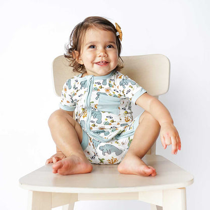 a happy baby sits on a wooden chair. she is wearing the "manatee" shortie. the print "manatee" unites the florida sea life in a majestic print. there is a variation of florida manatees, yellow and pink sea horses, yellow and pink starfish, bubbles, turtles, and coral patterned across a white background