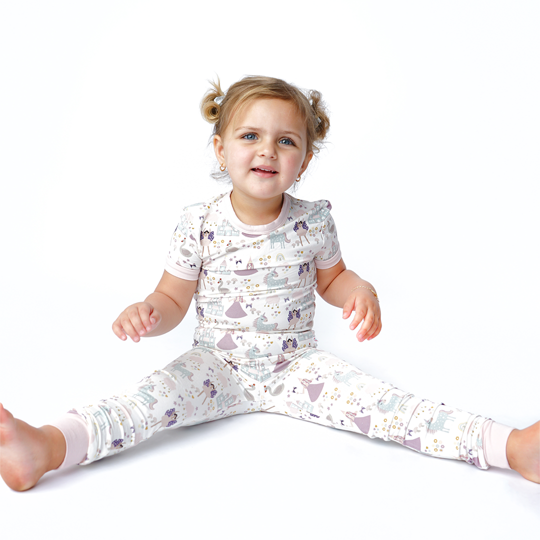 a girl sits on the floor with her legs out. she is wearing the "once upon a time" matching set. the "once upon a time" print is a mix of pinks, purples, whites, blues, and yellows. you can see fairy princess and regular princess, unicorns, swans, and castles. theres also hearts, stars and sparkles scattered around the print. 