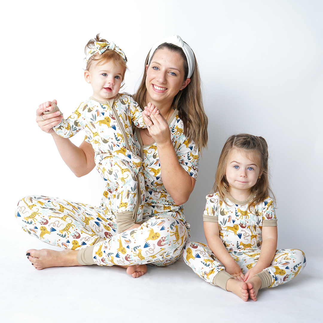 a mom and her 2 girls sit on the floor, all wearing their "jungle friends" matching apparel. the mom is in the women's top and jogger bottoms. she holds up the youngest daughter who is in the convertible and headband. the older of the 2 children sits next to them in the matching 2 piece pajama set. v