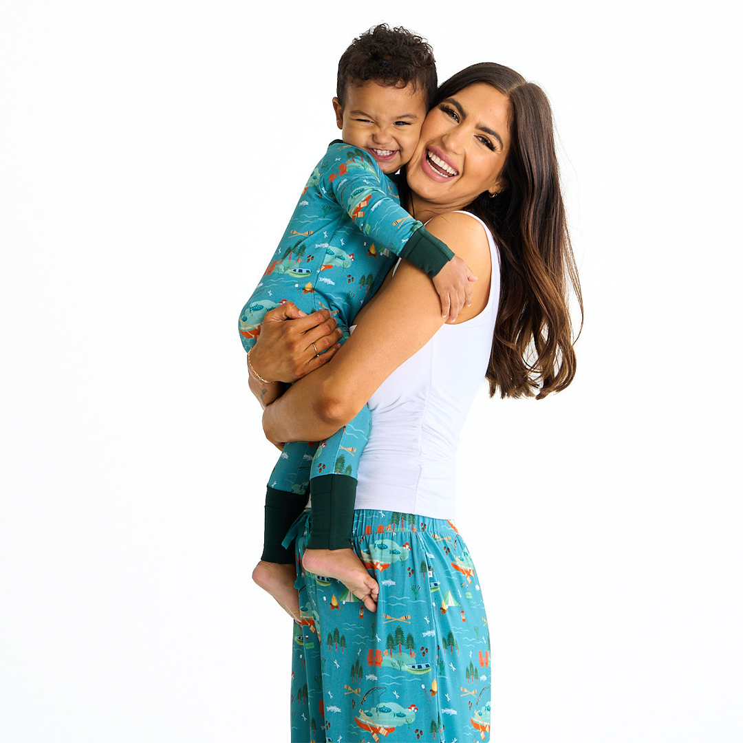 mom smiles and laughs into the camera she hugs her toddler boy. their faces are cutely smooshed together. the toddler wears the "jump in the lake" convertible. the mom wears the "jump in the lake" relaxed pants. the "jump in the lake" pattern is a print to capture the day in the life on a lake. you can fine fishing poles, tents, camp fires, adirondack chairs, marsh mellows, ores, canoes, forest trees, and different types of fish and fishing gear.