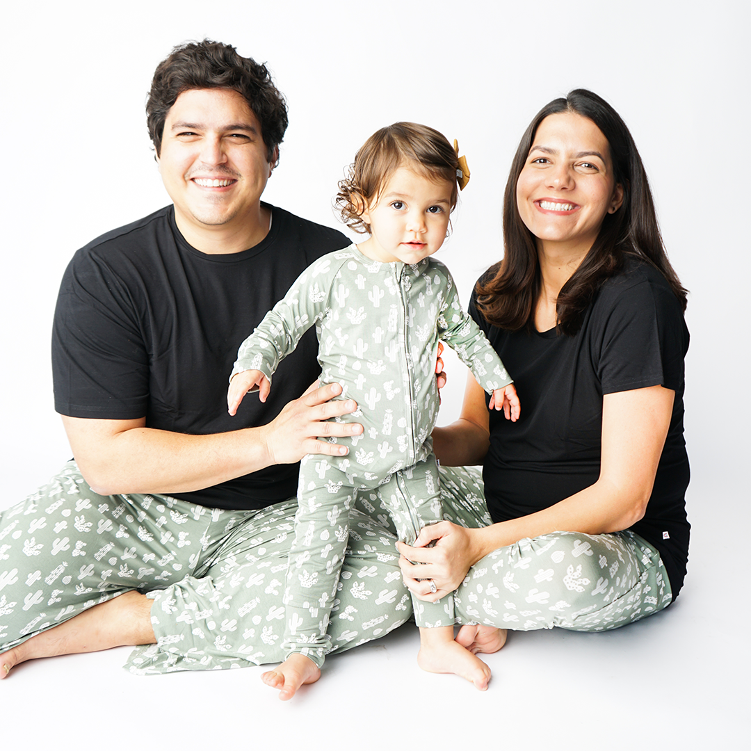 family of 3 smiles in their "stay sharp apparel. Mom and dad sit on the floor and hold on to their baby who stands. mom  is in the jogger pants, dad wears the relaxed pants, and the baby wears the convertible. the "stay sharp" print is a variety of different white cacti on a greyish/green background. 