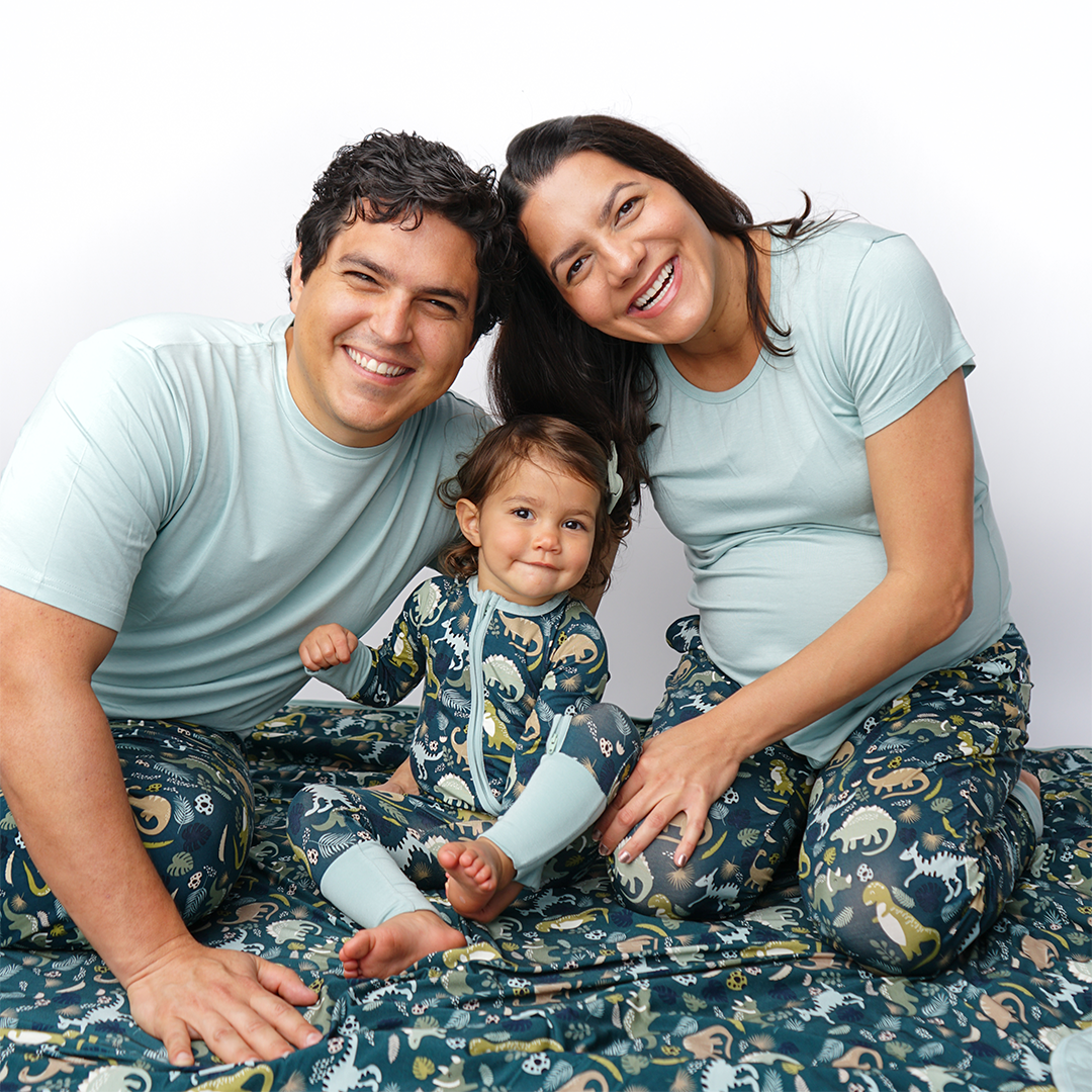 a couple and baby smile and pose for the camera. they are sitting on the "prehistoric friends" quilt. the dad and mom both wear "prehistoric friends" bottoms in the relaxed pants and the joggers. the baby wears the convertible. the "prehistoric friends" print is a mix of cute dinosaurs, eggs, and branches, all scattered on a dark green background.