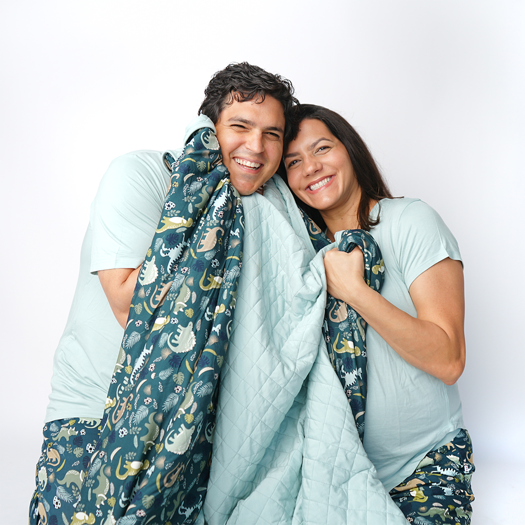 a couple poses and smiles for the camera. they are holding the "prehistoric friends" quilt. they are both wearing " prehistoric friends" prints in the relaxed pants and the joggers. the "prehistoric friends" print is a mix of cute dinosaurs, eggs, and branches, all scattered on a dark green background.